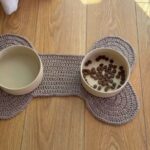 Taupe crochet dog bone placemat holding a water bowl and a food bowl, with the flat mat also shown on a wooden floor