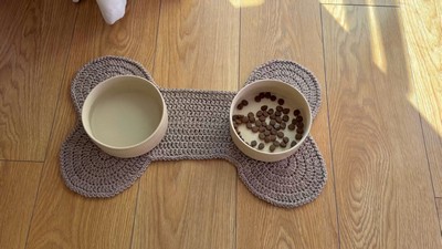 Taupe crochet dog bone placemat holding a water bowl and a food bowl, with the flat mat also shown on a wooden floor