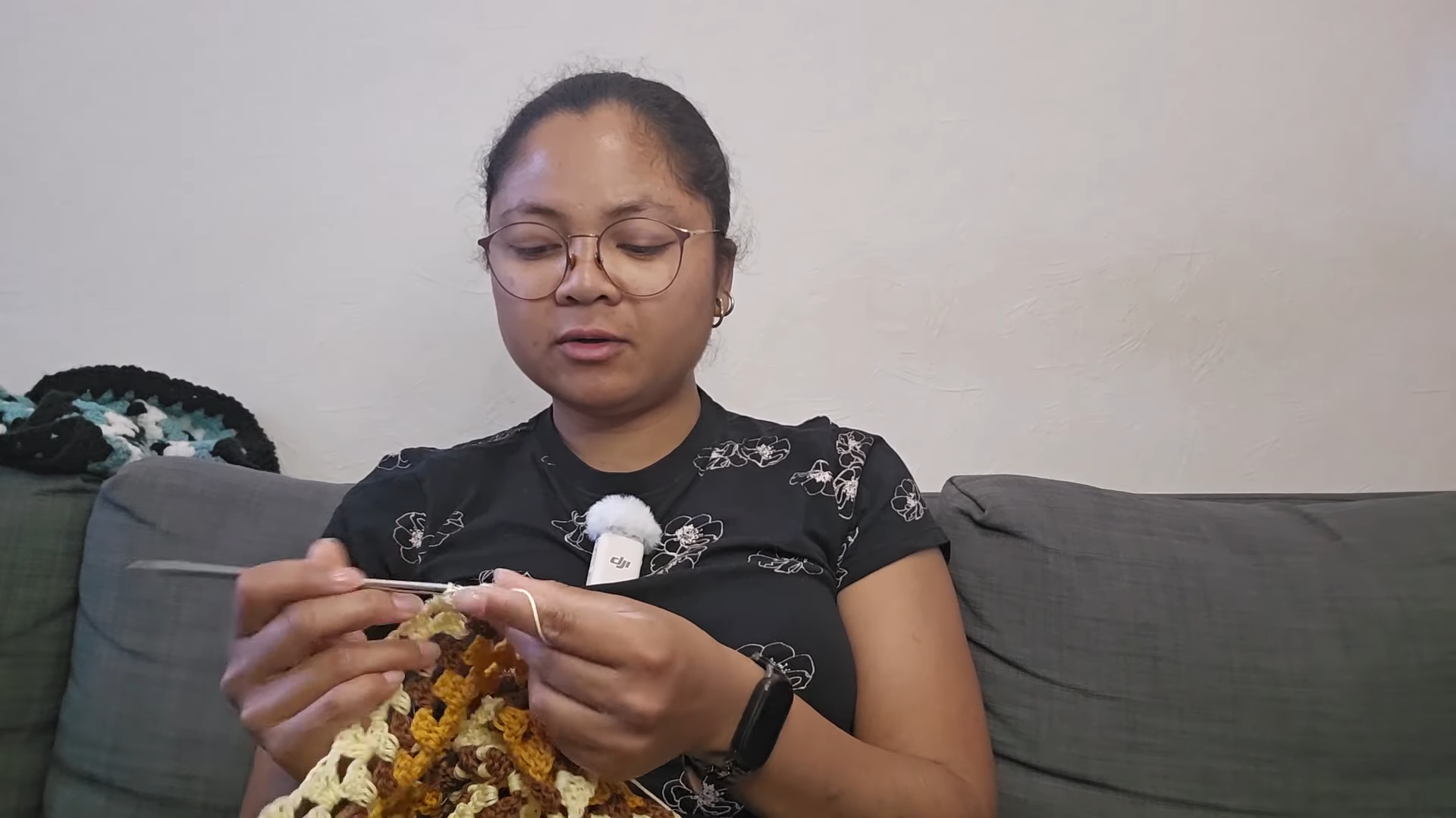 Woman crocheting a mustard and brown piece on a grey sofa.